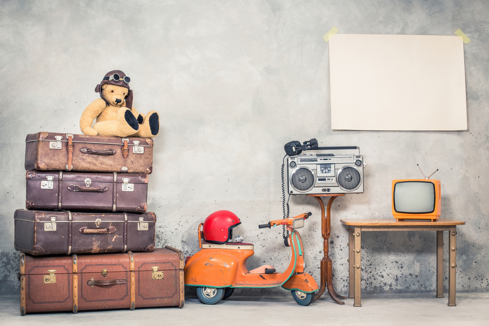 Retro Teddy Bear toy in aviator's hat, aged travel valises, children's pedal scooter, helmet, cassette boombox with headphones, TV, paper poster blank on concrete wall