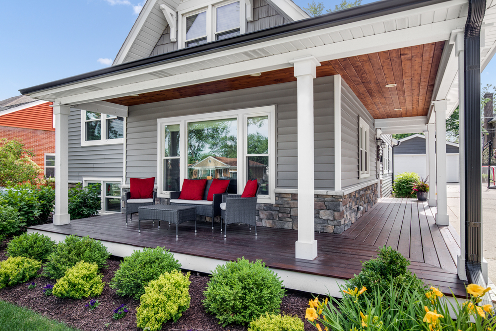 The exterior of a grey modern, suburban home with a covered porch and furniture featuring red accent pillows.