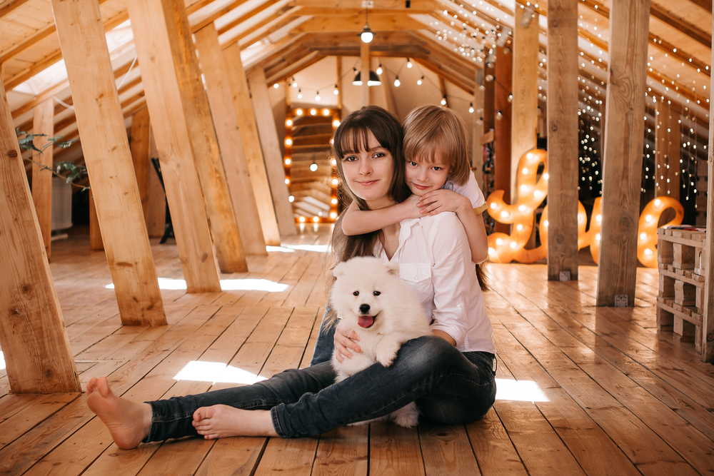 Young dark-haired mum with her son play with a Samoyed puppy in a wooden attic at home