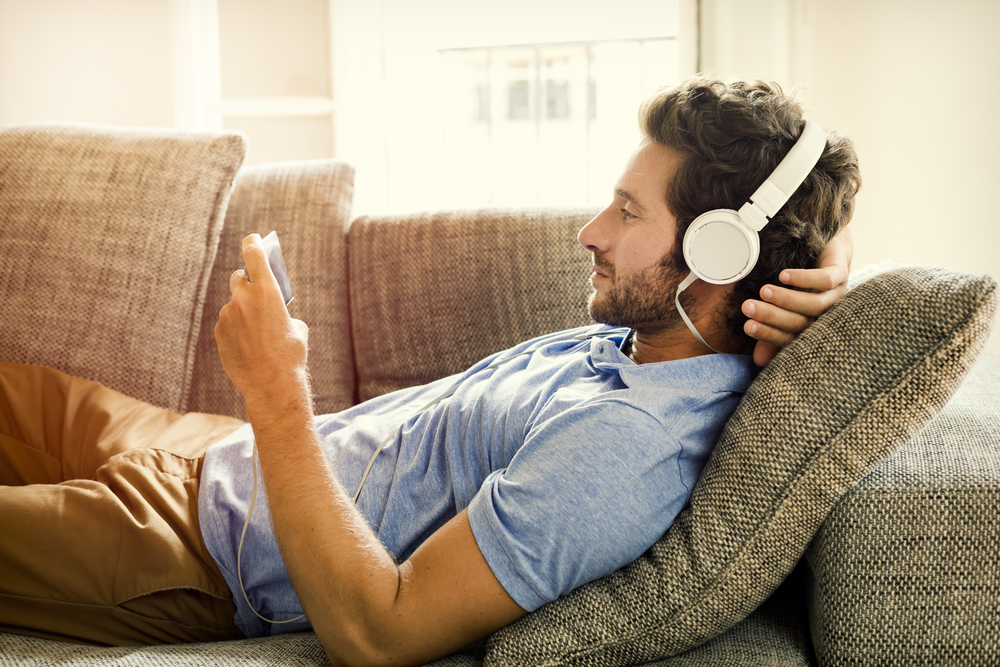 Man on couch, wearing headphones watches a movie on mobile phone