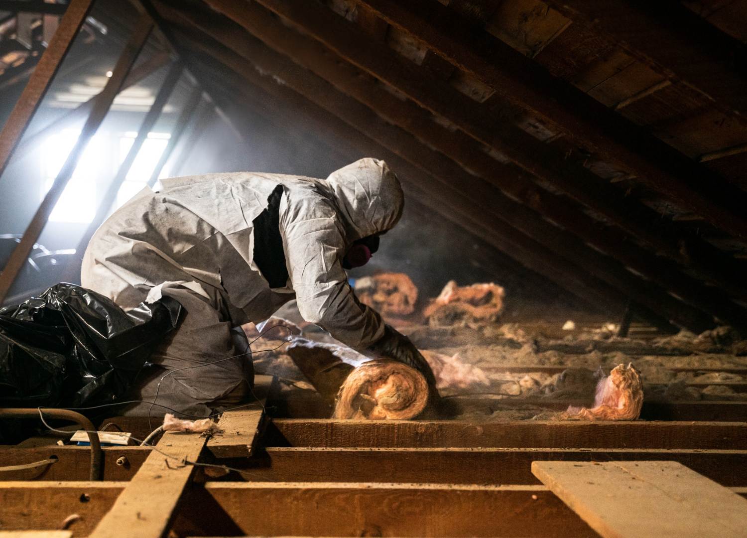 man in a white hazard suit and facemask removing old insulation from an attic.