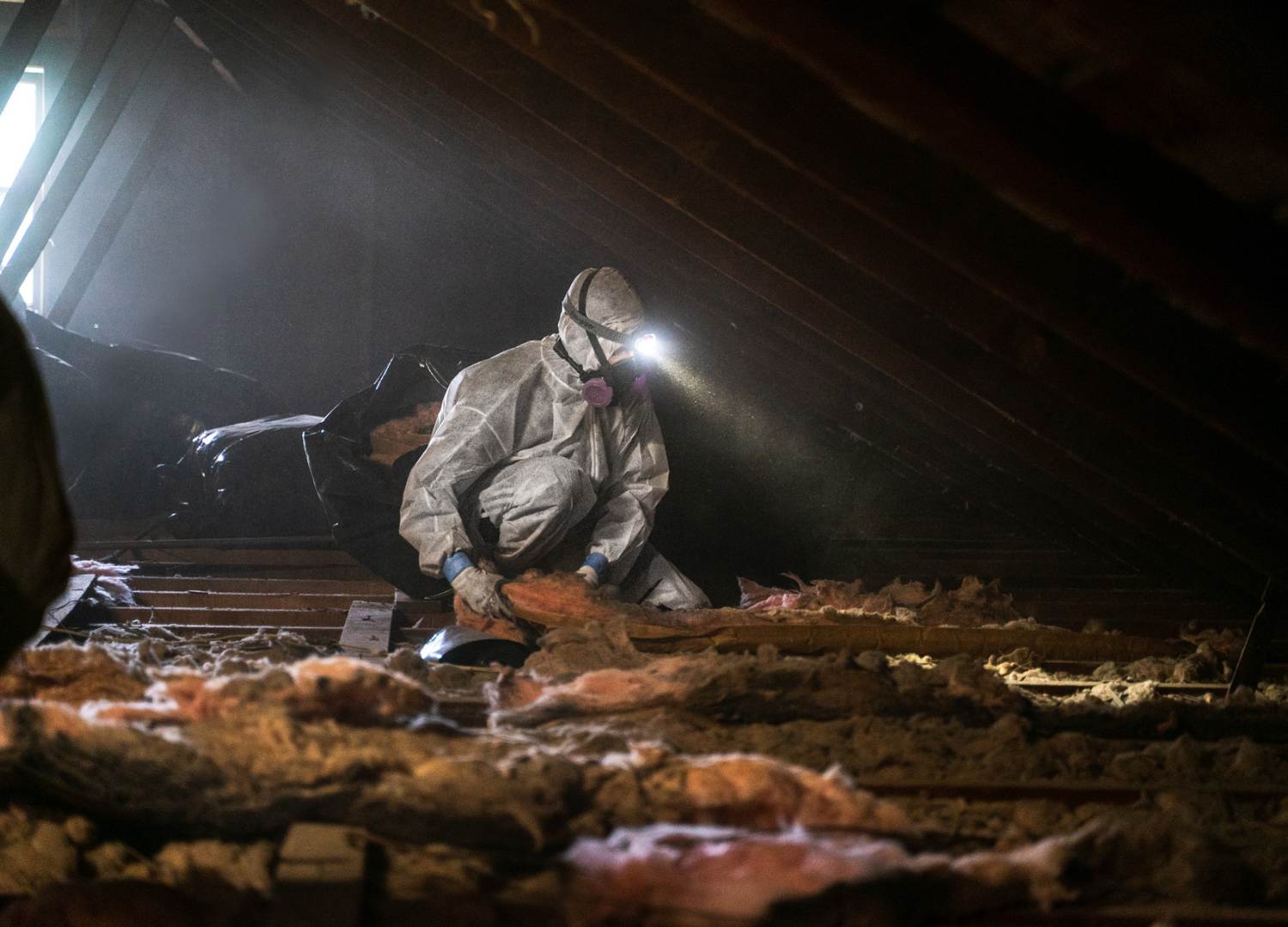 man in a white hazard suit and face mask removing old insulation in an attic.